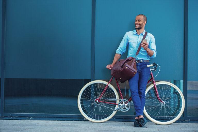 Full length portrait of handsome young Afro American man in casual clothes using phone, looking away and smiling while leaning on his bike, standing outdoors